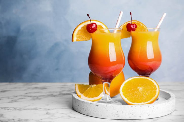 Zwei bunte Cocktails mit Orangen und Kirschen auf Marmorplatte vor blauem Hintergrund.