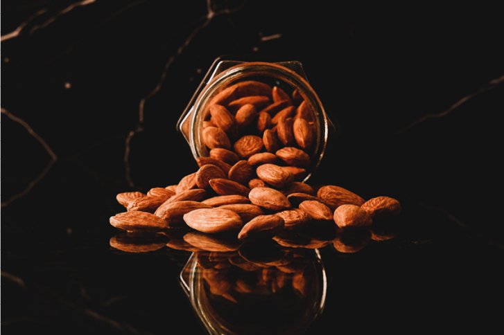 Ein Glasbehälter liegt, Mandeln ausstreuend, auf einer glänzenden, schwarzen Oberfläche.