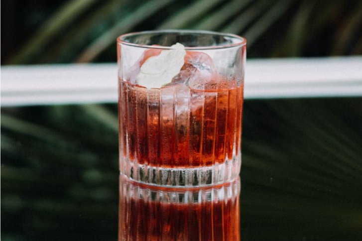Ein Glas mit rotem Getränk und Eiswürfeln steht auf einem reflektierenden Tisch.
