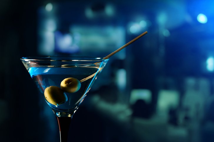 Martiniglas enthält zwei Oliven auf einem Spieß, mit verschwommenem, blau beleuchtetem Hintergrund.