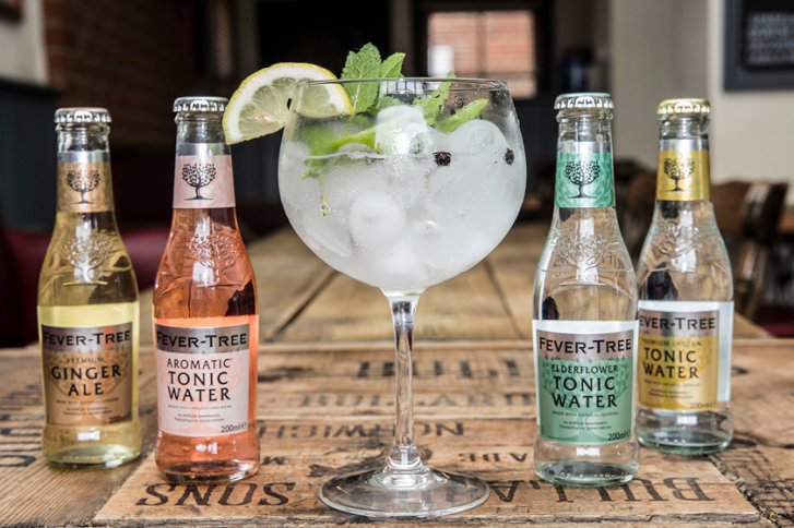 Glas steht auf Holztisch, gefüllt mit Eis, Zitrone, Minze, flankiert von vier Fever-Tree-Flaschen.