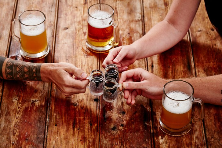 Hände stoßen mit Schnapsgläsern an, auf Holztisch mit zwei großen Bierkrügen im Hintergrund.