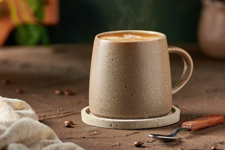 Keramiktasse dampft, auf Untersetzer; liegt auf braunem Tisch mit Kaffeebohnen und Löffel.