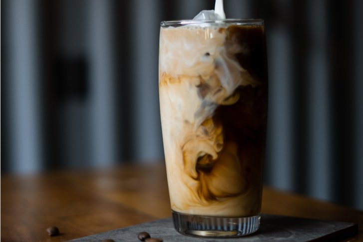 Ein Glas enthält sich vermischende Kaffee und Sahne, auf einem Holztisch in unscharfem Hintergrund.