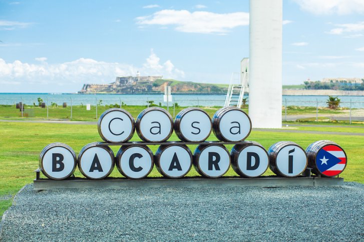 Fässer bilden Schrift "Casa Bacardi" vor Küstenlandschaft, Gras im Vordergrund, Meer im Hintergrund.