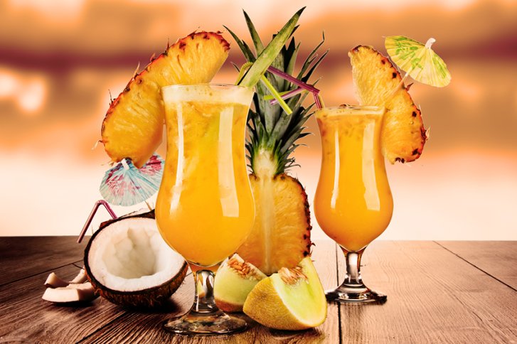 Zwei tropische Cocktails mit Ananas und Kokosnuss stehen auf einem Holztisch bei Sonnenuntergang.