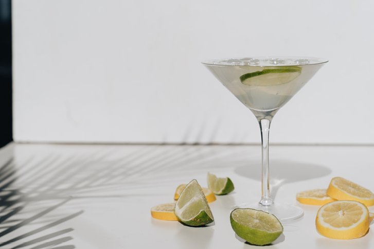 Cocktailglas steht auf Tisch mit Eiswürfeln und Limettenscheiben, umgeben von Zitrusscheiben.