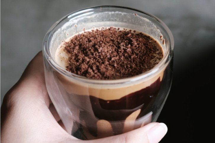 Hand hält Glas mit geschichtetem Dessert, bedeckt mit geraspelter Schokolade, in einem grauen Hintergrund.