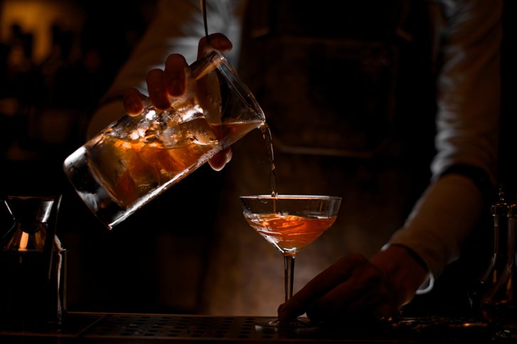Barkeeper gießt Getränk aus Mischglas in Cocktailglas, in dunkler Bar beleuchtet.