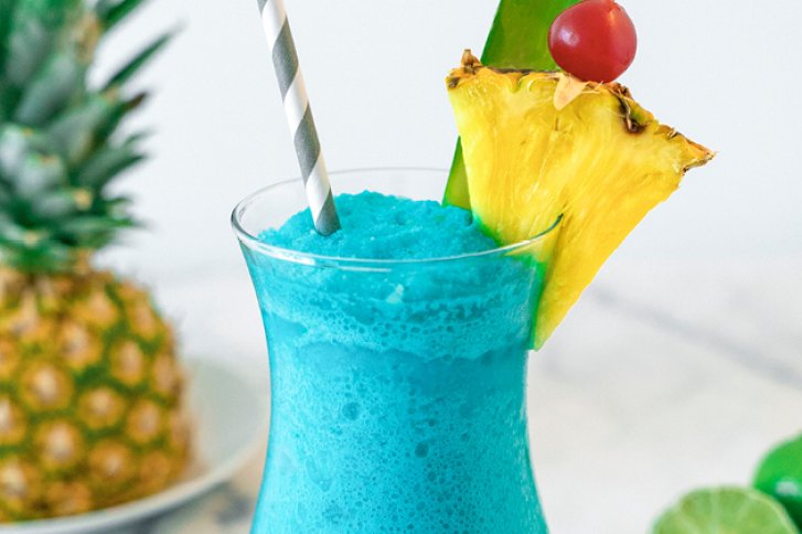 Ein blaues Getränk mit Strohhalm, garniert mit Ananas und Kirsche, vor Ananas.