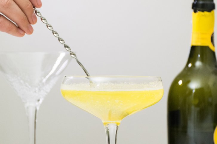 Eine Hand rührt mit einem Löffel in einem gelben Getränk, daneben steht eine Flasche.