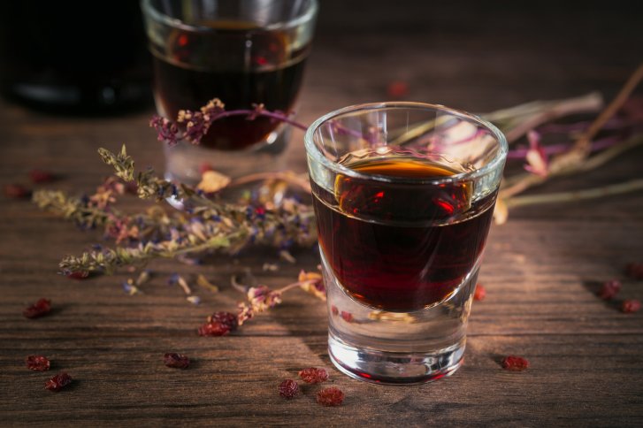 Ein Glas roter Likör steht auf Holztisch neben getrockneten Kräutern und Beeren.