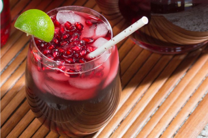 Ein Glas mit Granatapfelcocktail, garniert mit Limette, steht auf einem Holztisch in der Sonne.
