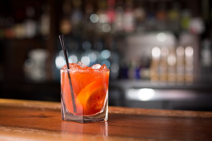 Glas mit rotem Getränk steht auf Holztresen, umgeben von unscharfem Bar-Hintergrund.