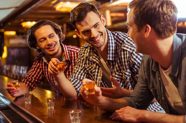 Drei Männer lachen, stoßen mit Schnapsgläsern an, in einer gemütlichen Bar.