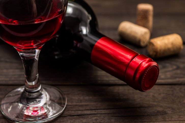 Rotweinglas steht auf Holztisch, neben liegender Flasche mit rotem Verschluss und Korken.