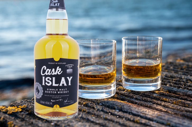 Flasche Cask Islay Whisky steht mit zwei gefüllten Gläsern auf Holztisch vor Wasser.