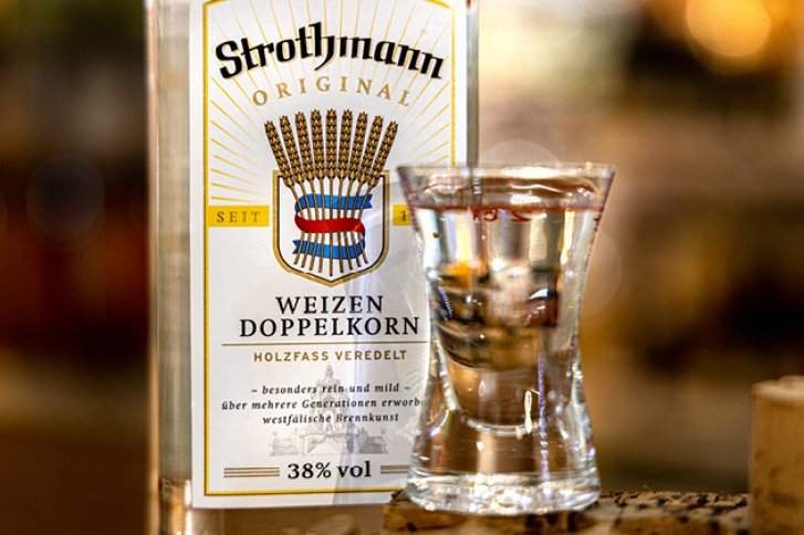 Ein Glas klarer Schnaps steht vor einer Strothmann Weizen Doppel­korn Flasche im Hintergrund.