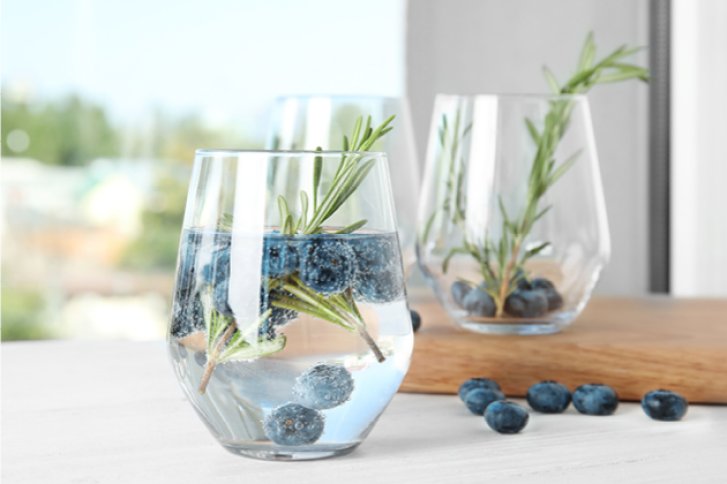 Glas mit Heidelbeeren und Rosmarin schwimmend, auf einem Holzbrett, helle Hintergrundszene.