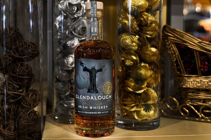 Flasche Whiskey steht ruhig zwischen dekorativen Glaszylindern mit goldenen und silbernen Blumen auf Holzregal.