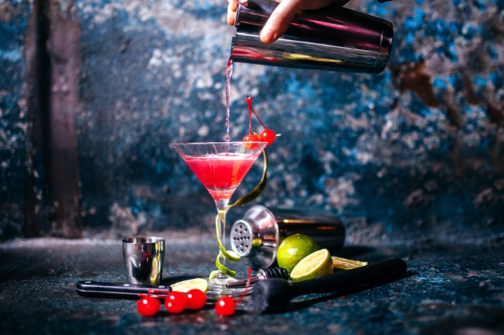 Cocktail im Martini-Glas, eingegossen von Shaker. Umgeben von Limetten, Kirschen, blauer Hintergrund.