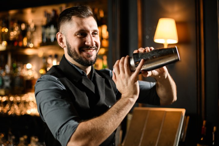 Ein Barkeeper schüttelt einen Cocktail-Shaker in einer stilvoll beleuchteten Bar-Umgebung.