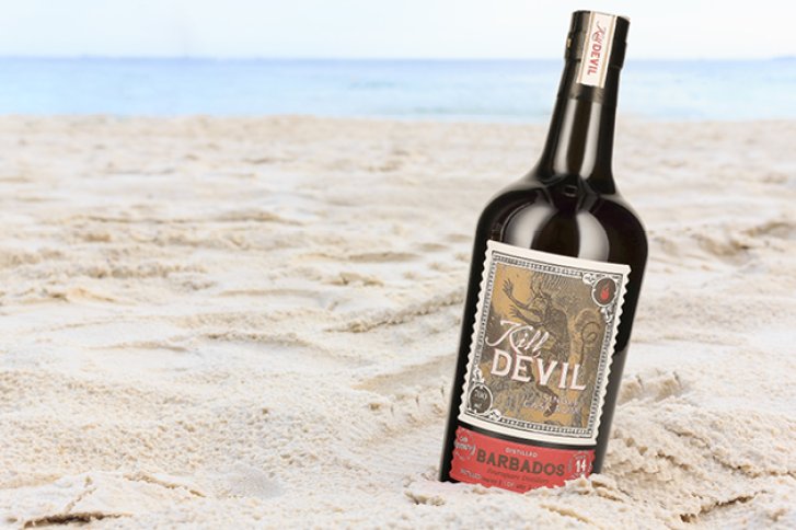 Eine Rumflasche steht schräg im Sand am Strand, im Hintergrund das Meer.