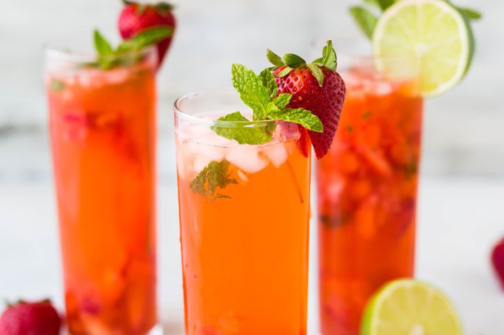 Drei Gläser mit Erdbeer-Getränken und Minzblättern stehen auf einem Tisch mit Limettenscheiben.