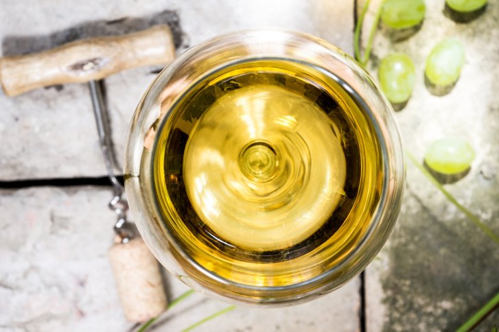 Gefülltes Weinglas stehend, umgeben von Korkenzieher und grünen Trauben, auf hellem Steinboden.