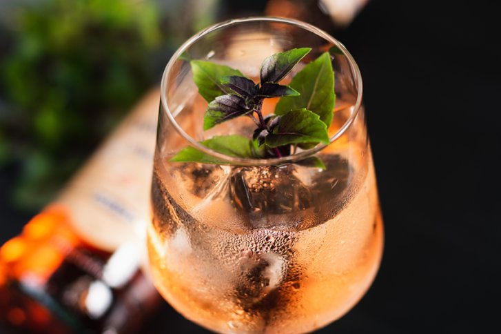 Ein Glas rosé Getränk mit Eiswürfeln und Basilikumblättern auf dunklem Hintergrund.