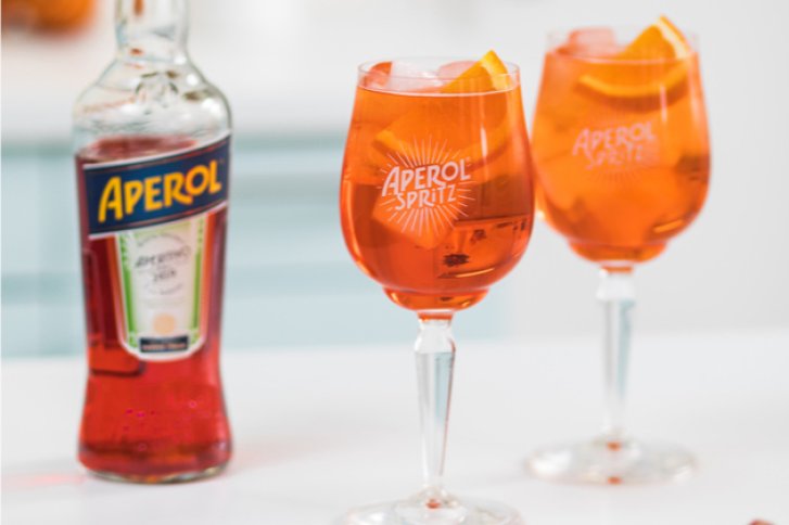 Zwei Aperol-Spritz-Gläser stehen auf einem Tisch, daneben eine Aperol-Flasche, helle Umgebung.