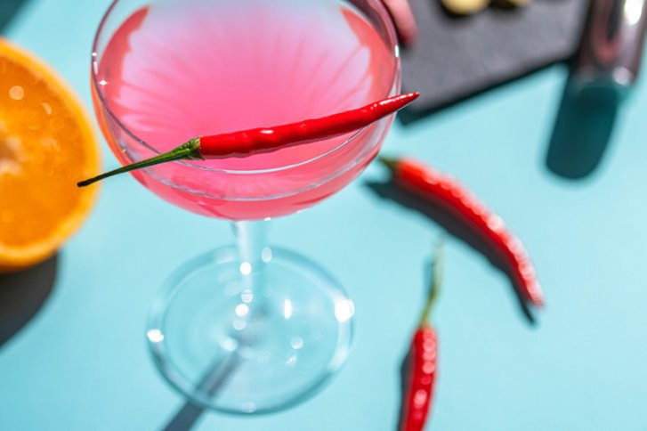 Rosa Cocktail im Glas mit roter Chilischote, umgeben von Orange und weiteren Chilis, auf blauem Hintergrund.