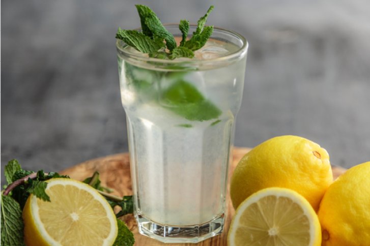 Ein Glas Zitronen-Minze-Getränk steht auf einem Holzbrett, umgeben von frischen Zitronen.