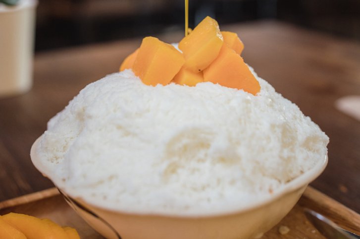 Schale mit Schnee-Eis; Mangostücke darauf; Sirup wird gegossen; Holzuntergrund im Hintergrund.