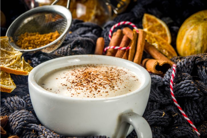 Weiße Tasse mit schaumigem Getränk, umgeben von Zimtstangen und getrockneten Orangen auf Wolle.
