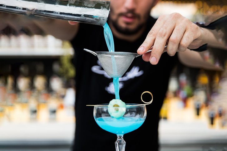 Blaue Flüssigkeit wird durch ein Sieb in ein Cocktailglas gegossen, Hintergrund unscharf.