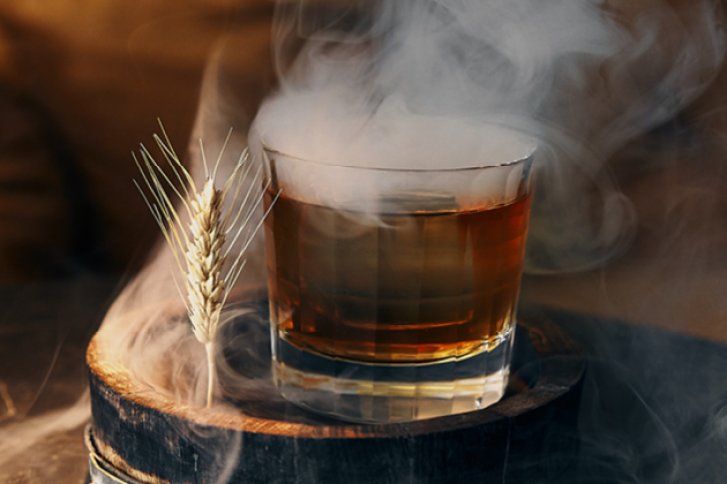 Ein Glas Whisky dampft auf einem Holzfass neben einem Weizenährenzweig in rauchiger Umgebung.