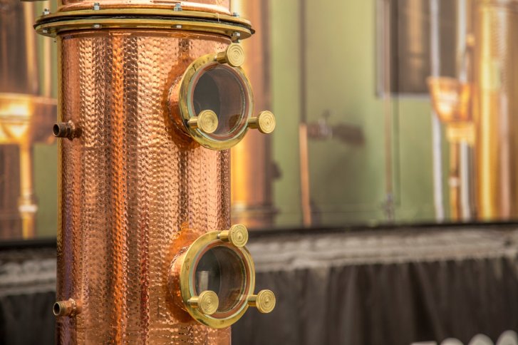 Kupferne Destillationsanlage mit runden Glasfenstern steht in industrieller Umgebung.