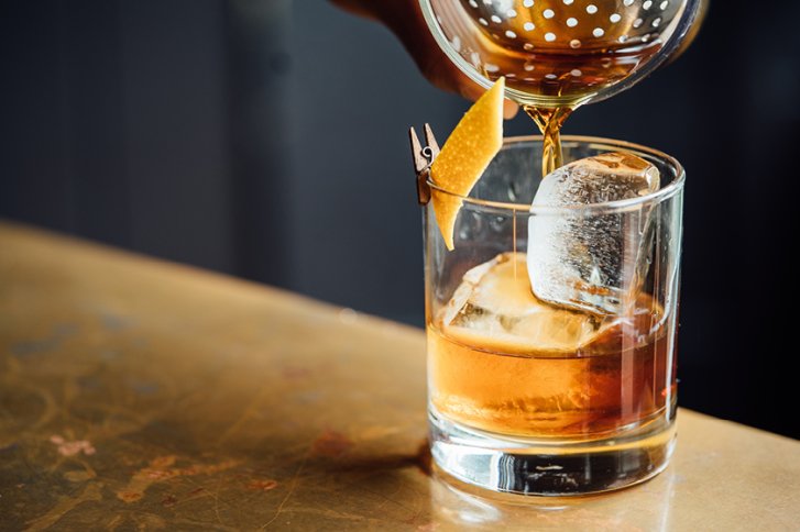 Ein Glas mit Eiswürfeln wird mit goldenem Getränk gefüllt, Zitronenschale dekoriert den Rand.