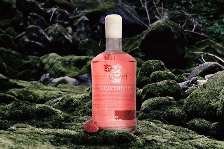 Eine Flasche Bärenman Dry Pink Gin steht auf moosbewachsenem Boden im Wald, Himbeere daneben.