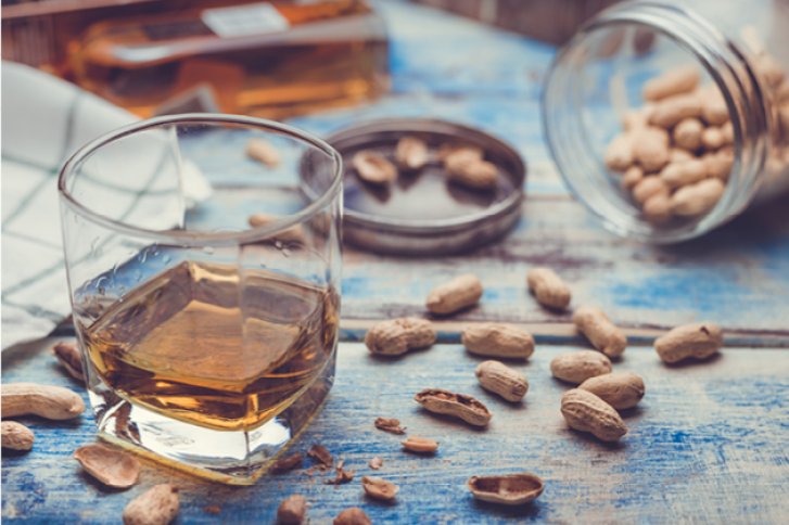 Glas mit Whisky steht auf blauem Holztisch, umgeben von Erdnüssen und offenem Glas.