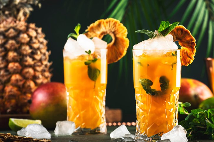 Zwei Cocktails mit Eis und Ananasdekoration stehen vor tropischen Früchten und Palmenblättern.