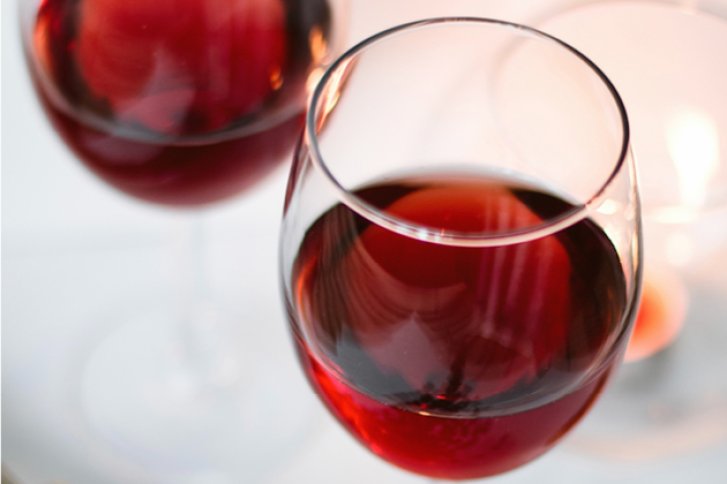 Rotweinglas – gefüllt und glänzend – auf hell gedecktem Tisch mit Kerzenlicht daneben.