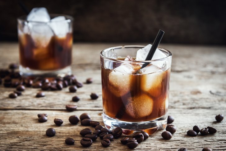 Ein Glas mit eisgekühltem dunklem Getränk steht auf einem rustikalen Holztisch, umgeben von Kaffeebohnen.