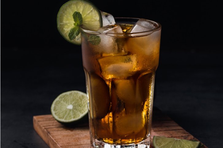 Ein Longdrinkglas mit Eis und Limettenscheiben steht auf einem Holzbrett, schwarzer Hintergrund.