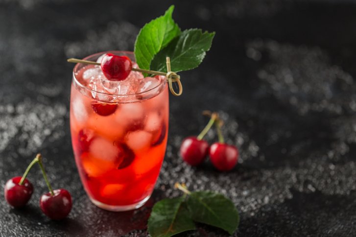 Ein Glas Cocktail mit Kirschen und Eiswürfeln steht auf einem dunklen Hintergrund.