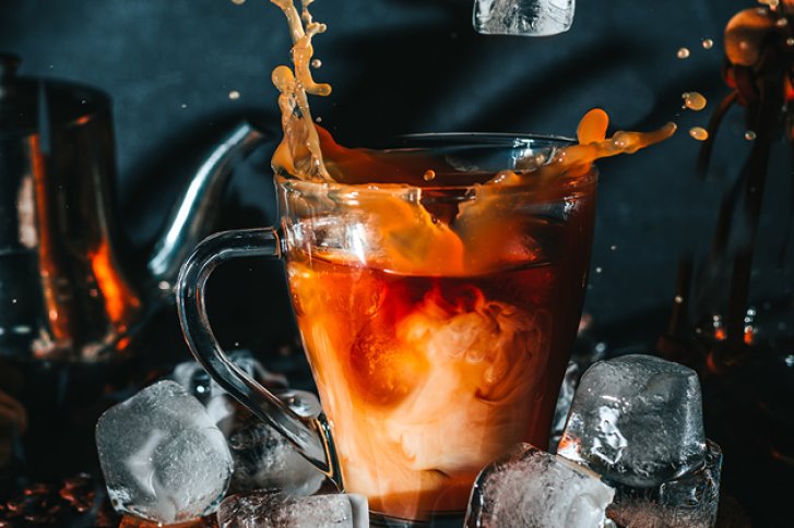 Tasse Kaffee wird mit Milch gesprenkelt, umgeben von Eiswürfeln auf dunklem Hintergrund.