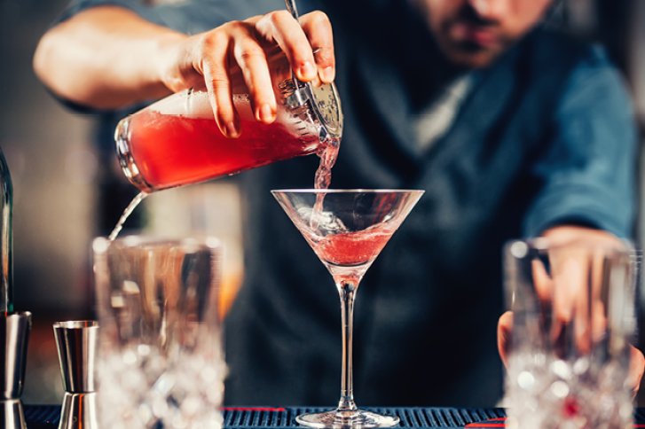 Ein Barkeeper gießt roten Cocktail in ein Martini-Glas, umgeben von Barzubehör.