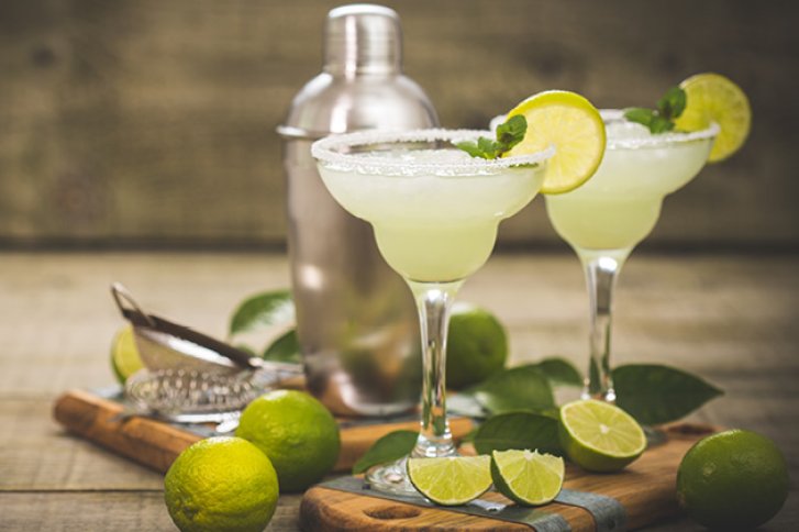 Zwei Margaritas mit Limettenscheiben stehen auf Holzbrett, umgeben von Limetten und Edelstahlshaker.