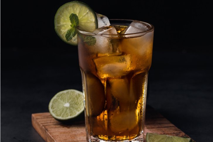 Ein Longdrinkglas mit Eiswürfeln und Limettenscheiben steht auf einem Holzbrett vor dunklem Hintergrund.